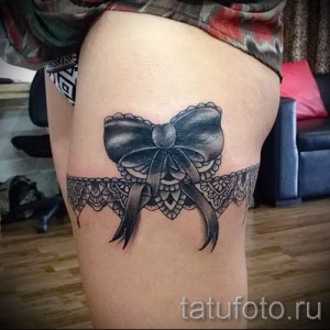 Фото тату: Черно-белая татуировка банта из кружева на бедре, выполненная в реалистичном стиле. Детализированный и изящный рисунок татуировки.