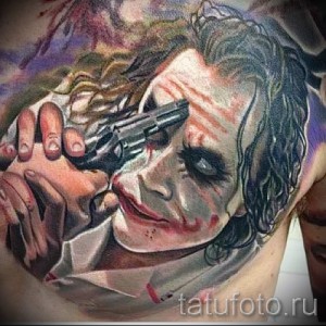 Фото тату Джокера с пистолетом на груди. Детализированное изображение лица злодея с безумным взглядом и оружием.
