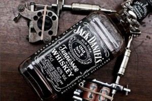Фото тату эскиза с изображением бутылки Jack Daniel's, тату-машинок и инструментов для татуировки на деревянной поверхности.