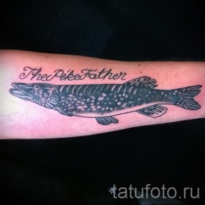 Фото тату щуки на руке с надписью "The Pike Father". Черно-серый стиль, реалистичная проработка рыбы и каллиграфическая надпись. Идеально для рыбаков и отцов.