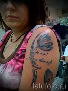 Фото тату тюльпана в серо-черных тонах на плече с надписью 'Life isn't a tiptoe through tulips'.