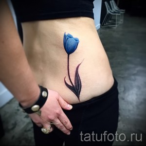 Фото тату синего тюльпана на женской талии. Элегантный рисунок татуировки в стиле минимализм, подчеркивающий изящество фигуры.