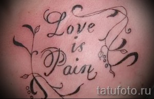 Фото тату на спине: надпись "Love is Pain" в каллиграфическом стиле, украшенная растительными элементами. Эмоциональный и выразительный рисунок татуировки.