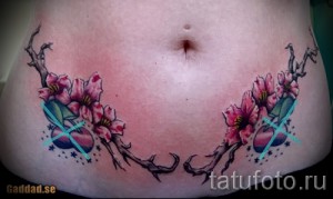Фото тату сакуры на животе. Изображение ветвей с розовыми цветами и синими акцентами. Оригинальный рисунок татуировки.