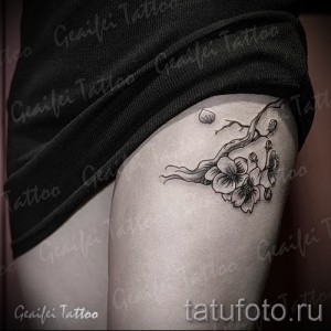 Фото тату: изящная черно-серая татуировка ветки сакуры с цветами и бутонами, расположенная на женской лопатке. Детализированный рисунок в стиле графика.