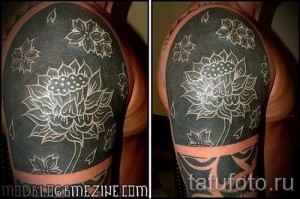 Фото татуировки на плече, выполненной в технике white ink tattoo, изображающей белые цветы лотоса на темном фоне.
