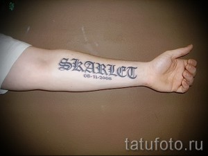 Фото тату имени 'Skarllet' и даты рождения '08.11.2008' на предплечье. Черно-белая татуировка, готический шрифт, памятный дизайн.