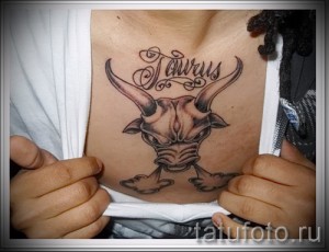 Фото татуировки Тельца с надписью "Taurus" на груди. Черно-серый рисунок с детальной проработкой головы быка.