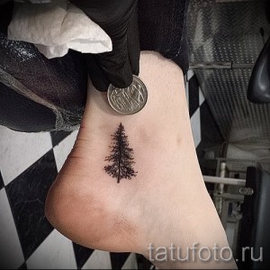 Фото миниатюрной татуировки на лодыжке, изображающей небольшую ель. Черно-белый рисунок, выполненный в технике тонких линий.