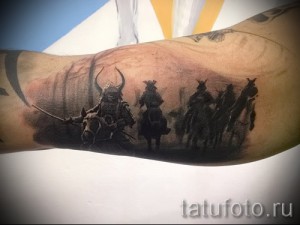 Фото тату на руке, изображающее группу воинов на лошадях в эпической битве. Черно-серый рисунок татуировки с детализированными фигурами и пейзажем.
