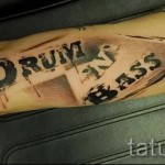Фото тату: надпись "Drum and Bass" в стиле граффити с элементами брызг и абстрактных форм на руке.
