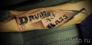 Фото тату: надпись "Drum and Bass" в стиле граффити с элементами брызг и абстрактных форм на руке.