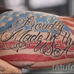 Фото тату: реалистичный рисунок флага США на бицепсе, с надписью 'Proudly Made in the USA'. Красный, белый и синий цвета.