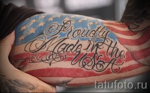 Фото тату: реалистичный рисунок флага США на бицепсе, с надписью 'Proudly Made in the USA'. Красный, белый и синий цвета.