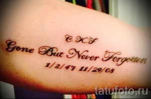 Фото тату на руке: надпись "Gone But Never Forgotten" с датами, выполненная каллиграфическим шрифтом в красных тонах.