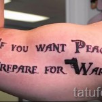 Фото тату: фраза "If you want peace prepare for war" с изображением пистолета на предплечье, выполненная в классическом стиле леттеринга.