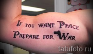 Фото тату: фраза "If you want peace prepare for war" с изображением пистолета на предплечье, выполненная в классическом стиле леттеринга.