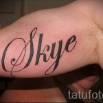 Фото тату: изящная надпись "Skye" на бицепсе, выполненная черными чернилами в каллиграфическом стиле с тонкими линиями и завитушками.