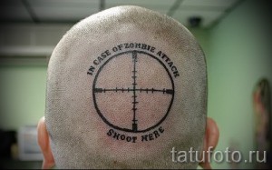 Фото татуировки на голове, изображающей прицел с надписью "In case of zombie attack shoot here", выполненной в черном цвете.