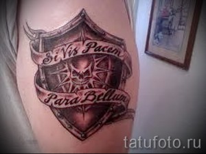 Фото тату на бедре: щит с изображением черепа и надписью 'Si Vis Pacem Para Bellum', выполненный в реалистичном стиле с использованием черных и серых оттенков.