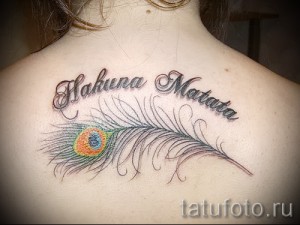 Фото тату: надпись 'Hakuna Matata' и изображение пера павлина на спине девушки. Перо выполнено в ярких цветах, а надпись – в черном цвете.