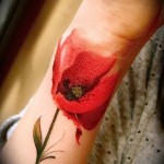Фото тату: акварельный рисунок красного мака на запястье. Насыщенный красный цвет, тонкие линии стебля и листьев, создающие эффект акварели.