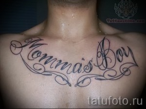 Фото тату надписи 'Momma's Boy' на груди, выполненной в каллиграфическом стиле с тонкими завитушками.