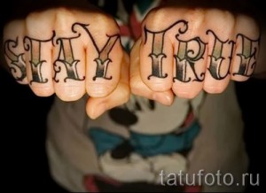 Фото тату с надписью 'Stay True' на костяшках пальцев, выполненной черными чернилами в классическом стиле. Идеально для любителей минимализма и мотивационных татуировок.