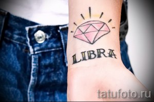 Фото тату на запястье: розовый бриллиант с надписью LIBRA. Эскиз татуировки выполнен в акварельном стиле с использованием нежных оттенков.