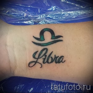 Фото тату: татуировка Весы с надписью 'Libra' на спине, выполненная в черно-зеленой гамме с использованием плавных линий и градиентов.