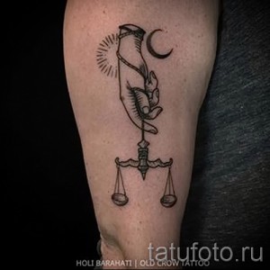 Фото тату Весы на руке. Изображение выполнено в черном цвете, с использованием тонких линий. Весы держат в руке, символизирующей контроль и ответственность.