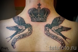 Фото татуировки на спине: корона в окружении двух ласточек с лентами, выполненная в черно-серых тонах.