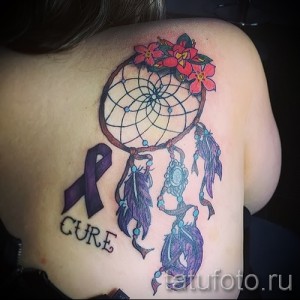 Фото татуировки ловца снов на спине с добавлением фиолетовой ленты поддержки и надписи 'CURE'. Детализированный рисунок татуировки с использованием различных оттенков.