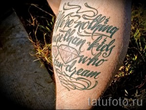 Фото тату с надписью 'We're nothing more than kids who can dream' и изображением бриллианта, выполненного в черно-серых тонах. Детализированный рисунок татуировки на ноге.