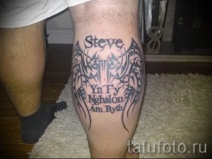 Фото тату на голени с именем "Steve", символами и крыльями. Черно-белое изображение, детализированная проработка и готический стиль.