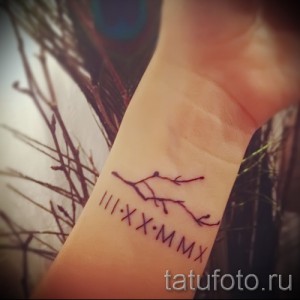 Фото тату с изображением ветки дерева и даты, написанной римскими цифрами, на запястье. Рисунок татуировки выполнен в тонких линиях, выглядит нежно и элегантно.