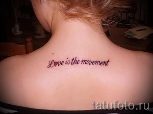 Фото тату надписи "Love is the movement" на спине девушки. Красная надпись с маленьким сердечком, выполненная изящным шрифтом.