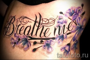 Фото тату надписи 'Breathe me' с изображением фиолетовых цветов на боку. Элегантный шрифт и яркие краски.