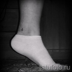 Фото тату: крошечный черный перевернутый крест на лодыжке девушки в белых носках.