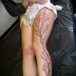 Фото тату ветви сакуры на бедре. Тонкие черные ветви с нежно-розовыми цветами, выполненные в реалистичном стиле, создают изящный и женственный образ.