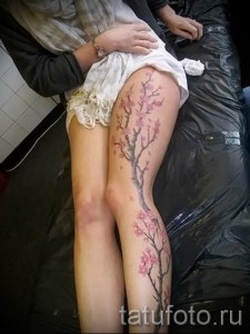 Фото тату ветви сакуры на бедре. Тонкие черные ветви с нежно-розовыми цветами, выполненные в реалистичном стиле, создают изящный и женственный образ.