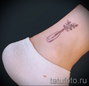 Фото тату с изображением небольшой бутылки, из которой растут цветы, на лодыжке. Рисунок выполнен в минималистичном стиле с использованием тонких линий.