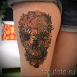Фото тату с изображением черепа, полностью покрытого цветами. Красивый рисунок татуировки на бедре с использованием теплых и ярких оттенков.