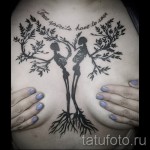 Фото татуировки на спине девушки с изображением двух скелетов, обнимающихся под деревом жизни, с надписью на английском языке.