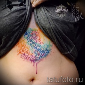 Фото акварельной тату 'Цветок жизни' с яркими радужными цветами, расположенной на животе девушки.
