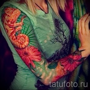 Фото тату хамелеона на руке, выполненного в ярких красных и оранжевых тонах. Рисунок изображает хамелеона, обвивающего руку среди цветущих растений.