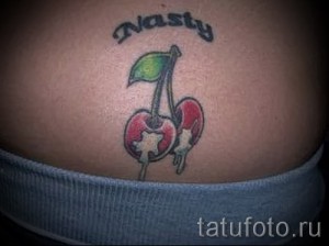 Фото тату двух вишен с зеленым листком и надписью 'Nasty' на коже. Яркий, провокационный рисунок татуировки.