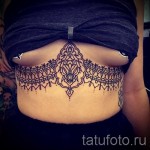 Фото тату: изящная черно-белая татуировка, имитирующая кружево, расположена под грудью. Детализированный рисунок с тонкими линиями и узорами.