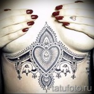 Фото тату: сложный узор в стиле мандалы, расположенный под грудью. Черно-белая татуировка с геометрическими элементами и тонкими линиями.