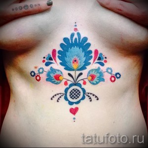 Фото тату: яркий рисунок в стиле folk art, изображающий цветы и птиц, расположенный между грудью. Детализированная татуировка с использованием ярких цветов.